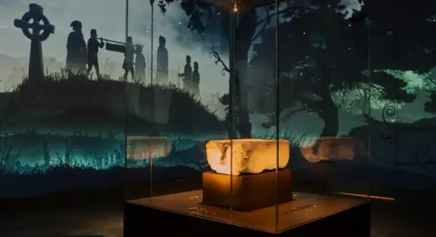 The Stone of Destiny sits in a glass case with a projected background showing silhouetted figures and a cross