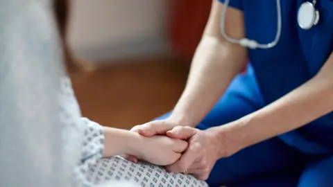 Phil Boorman via Getty Images A clinician wearing a blue uniform clasps the hands of a patient wearing a patterned hospital gown. 