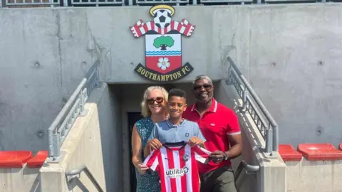 Nicky Ajanaku Kayo is stood holding a Southampton FC shirt with his parents behind him at a football stadium