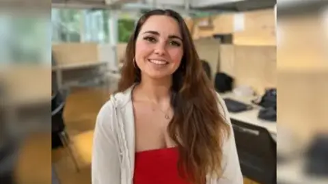 The image shows a girl with long brown hair smiling; she is also wearing a red top and white cardigan. Behind her desks and chairs can be seen.