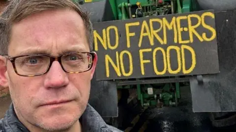 Daryl Corps Councillor Daryl Corps pictured in front of a sign that reads "no farmers no food" in yellow capital letters. He is looking directly at the camera and frowning slightly.