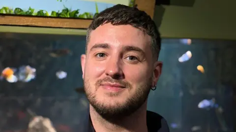 A young man with a beard and earring in front of an aquarium tank of blue tropical fish