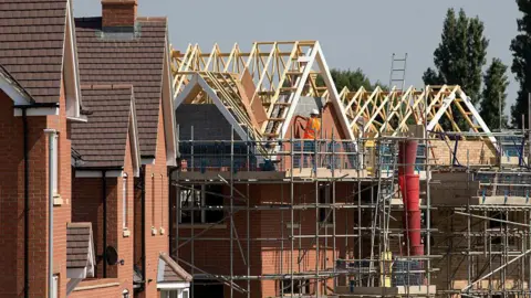 A row of partially built red-brick homes. There is a construction worker standing on a scaffolding platform next to exposed roof trusses.
