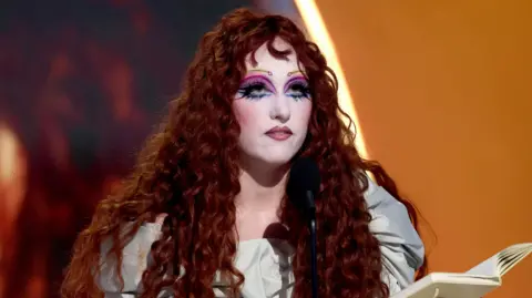 Chappell Roan during her acceptance speech for Best New Artist at the Grammy Awards. Her curly red hair is worn loose and she wears elaborate makeup including pencil thin brows and pink, orange and blue eyeshadow. She wears a silver dress, holding a notebook in her left hand and her trophy in her right. 