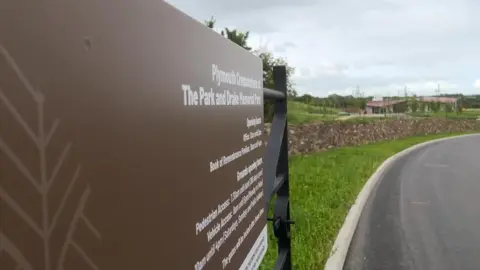 A picture of the entrance to the crematorium. The gate has a brown sign containing white writing. The crematorium can be seen in the background surrounded by grass.