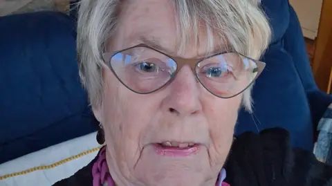 Jayne Brearley A woman with short grey hair and glasses is looking at the camera in this selfie.