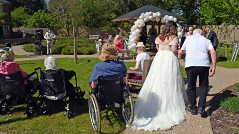 Emma Noakes Emma in her wedding dress is accompanied by George in a white top as they walk down a path. On the left, people in wheelchairs can be seen looking at a gazebo with a balloon arch formed at the entrance.