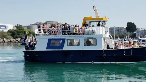 Passengers waving while standing aboard a vessel run by Plymouth Boat Trips in Plymouth Sound,