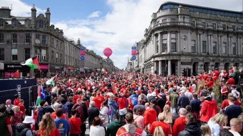 PA Media A large crowd of football fans wearing red in front of granite buildings and a blue sky,