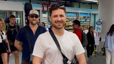 Handout Mr Eury, pictured in what appears to be an airport. He is wearing a white T-shirt with a black cross-body bag strap, and glasses on his head. He has very dark curly hair and dark stubble. 