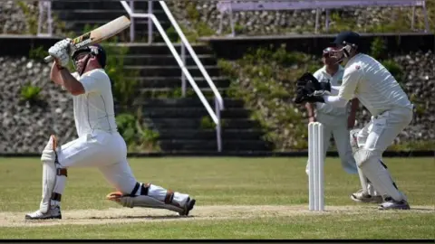 A batsman stretches out of his crease to hit a scoring shot. A wicket keeper stands behind the stumps. Both men are in full whites cricket gear. Another fielder is seen in the background. The pitch they are playing on is well maintained.