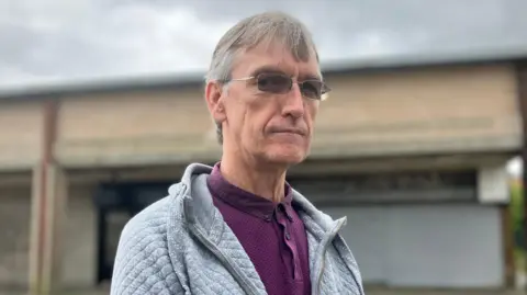 David Taylor wearing a purple top and grey hoodie. He has grey hair and is wearing square metal rimmed glasses.  