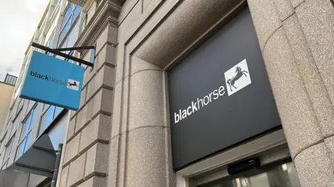 A black sign with white writing which says 'black horse', next to an image of a black horse on a white background. The sign is positioned above a door, and is surrounded by granite brickwork. To the left of the image is a light blue sign which also says "black horse".