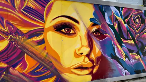 7th Pencil A painting on a wall of a woman's face in yellow surrounded by purple flowers.