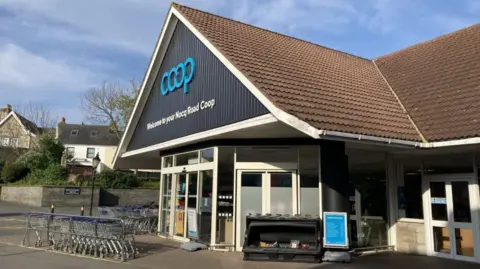 A CI Coop store in Guernsey. The building features a triangular roof with brown tiling and the organisation's logo in blue at the top of the front façade. 