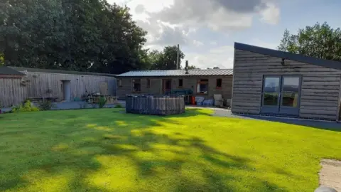 Cheshire East Council A wooden structure is built at the end of a garden area. The structure fills the width of the garden. There is a large glass door entrance on the right and windows lining the structure on the left. 