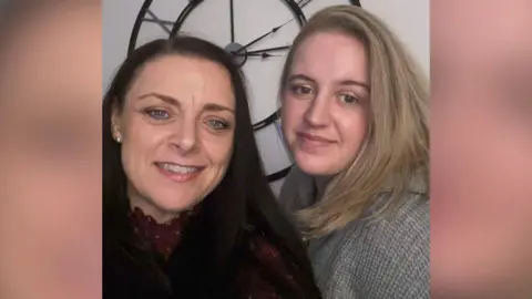 Family photo Lisa Wolff and her daughter Abbigail Smith on her left hand side. Lisa has long dark hair and is wearing a dark red top. Abbi has long blonde hair and is wearing a grey jumper. They are standing in front of a white wall with a black metal clock.
