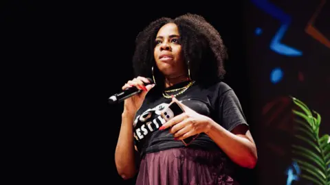 Black British Book Festival Selina Brown, with curly black hair to her shoulders, holds a microphone as she stands on stage, wearing a black top with white writing and a burgundy skirt.