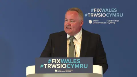Darren Millar at a podium at Welsh Conservative conference, which has the words #FIXWALES and #TRWSIOCYMRU with Tory branding. He is swearing a black suit, green tie and white shirt.