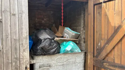 Ben Schofield/BBC A shot inside a communal bin store near Louise's flat. A large metal bin is overflowing with full refuse sacks, on top of which are empty cardboard boxes. The brick structure of the bin store can be seen behind the bin, though the inside of the store is dark. On the left of the image, part of one wooden door is visible, still in its closed position, while on the right the other wooden door hangs open.