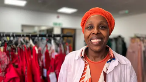Florence Okello stands in front of a rack of red dresses. She is wearing an orange scarf on her head and brightly coloured clothes. She is smiling at the camera.