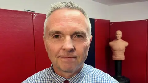 Iain Ramsay staring straight at the camera. He has short, grey hair and is wearing a blue and white checked shirt. He is standing in front of a wall with red mats on it and an armless mannequin is in the background.