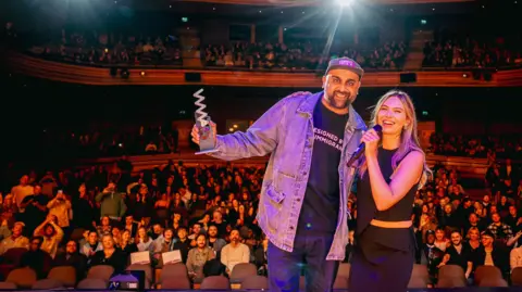 Welsh Music Prize Aly Jamal or Don Leisure pictured on stage at the Wales Millennium Centre, with the audience applauding behind, receiving his award from an unnamed female presenter.
