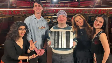 Supplied A group of five people standing side by side and smiling at the camera. Left to right, a woman with long black hair and glasses, and a black top, points with both hands at the central figure holding an award; a taller man with short dark dark hair and pale blue shirt; a person with a lilac beanie hat and stripy blue and cream jumper holding a sheet of silver designed as an open book; a woman with red hair  and a black dress, and a woman with long dark hair and a black dress.