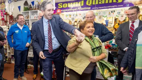 Fine Gael/PA Taoiseach Simon Harris, wearing a navy suit, blue shirt and check red tie, smiling and holding hands with a shorter woman with short light brown hair. She is also similar and is wearing dark leggings, a cream shirt and a green silk scarf. They are surrounded by five smiling people. They are standing inside a gift shop. 