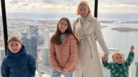 Rebecca wearing a cream belted coat, with her three children Noah (left), Leila (centre) and Artie right), standing at a large window at the top of a high-rise building overlooking a cityscape. Noah is wearing a dark blue puffer jacket, Leila is wearing a pink coat and Artie is wearing a green puffer coat with what look like cartoon tigers on them. 