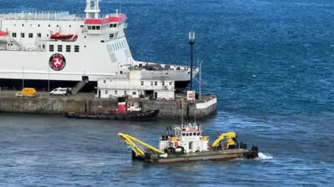 A small dredging boat is in Douglas Harbour. It has a small yellow crane-like device at one end and another piece of yellow equipment at the other. The Manxman ferry is visible on the other side of the pier.