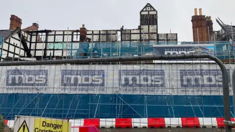 BBC A large black and white Tudor building, with sections of the site discoloured from a fire and holes in the wall. 