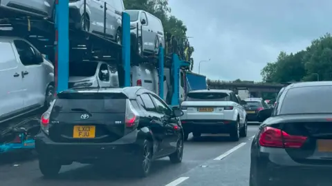 BBC A queue of mainly black and white cars and a car transporter on a motorway