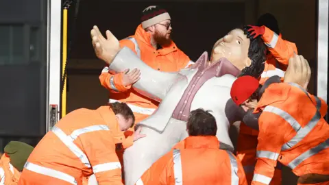Five men in high viz orange jackets, carrying one of Beryl Cooks sculptures out of a van. The sculpture is of 'Dancing Tom'.