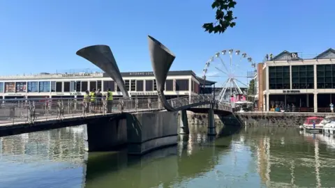Peros' Bridge in Bristol Harbour, with the nearer section visibly higher than the far section. Workmen in high vis jackets are standing on the bridge.