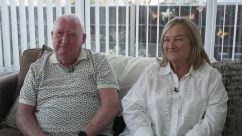 A man with white hair, wearing a patterned top sits on a sofa next to a woman with blonde hair, wearing a white blouse. They are smiling at the camera and are sitting in a conservatory. 