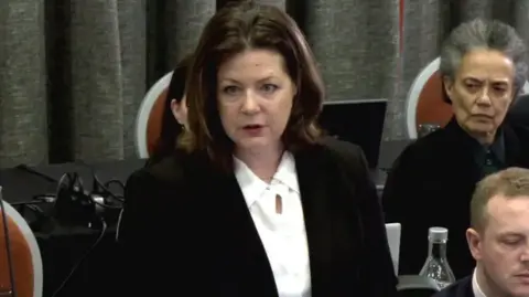 Brenda Campbell KC has shoulder length brown hair and is wearing a white shirt and black blazer. She is speaking in the picture.