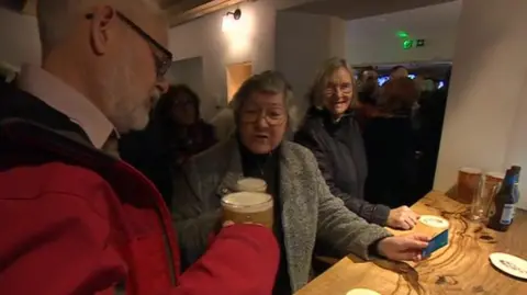 Two people with pints of beer at a bar, doing a 'cheers' motion, having reopened the pub.