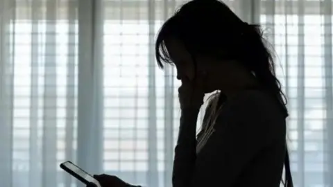 Getty Images Silhouette of a woman with long hair with her right hand on the side of her face and her left hand clutching a mobile phone, which she is looking at. There is a grey vertical blind in the background.
