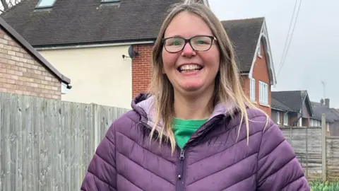 Julia Gregory/BBC Close up shot of Annette Whymark smiling to camera standing against a fence with houses in the background