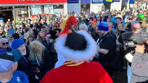 Crowds of fans in Cinderford town centre, with a couple of TV cameras also filming the band as they interact with members of the public.