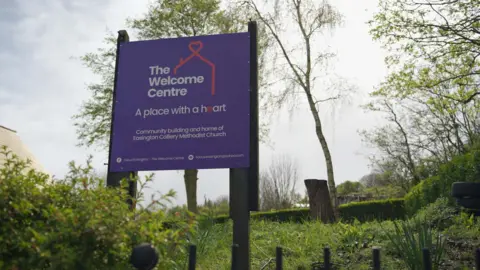 Grassed area with a purple sign on two posts reading "The Welcome Centre - A Place with a Heart". Behind it are shrubs and a two trees