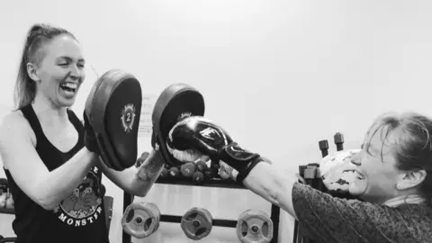 A black and white picture of two women practicing boxing. Both women are smiling and have their hair up. The woman on the left is wearing a black tank top and punching mitts. The woman on the right is wearing a top and boxing gloves. The photo is them from the waist up. Behind them is a blank wall and some weights. 