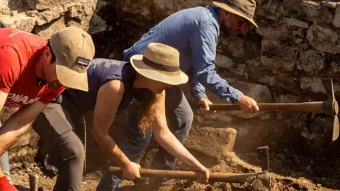 Alexander Jansen/Durham University Two men and a woman digging in the sunshine at the Auckland Palace excavation site. They are wearing hats and leaning as dust flies in the air.