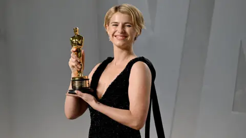 Jessie Buckley with short blonde hair holds up a gold Oscar award with both hands while wearing a black, sleeveless dress
