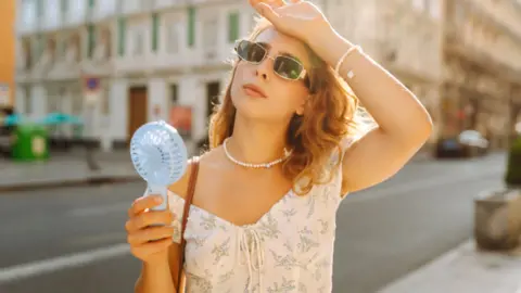 Woman with blonde hair, wearing sunglasses has her left hand on her forehead and a small electric fan in her right hand.