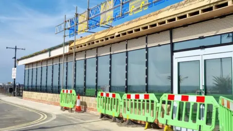 Isle of Wight Council A one-storey building made up of glass windows. In front of the building are green building work barriers.