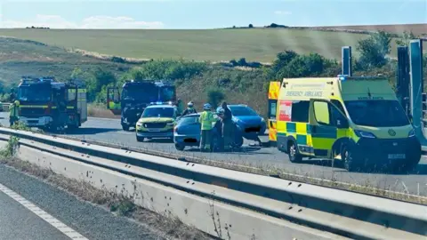 Emergency service vehicles near to a couple of cars on an A road. There are green hills in the background.