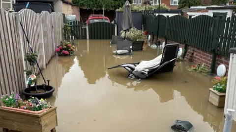 Lisa Daniels A residential back garden that has been extensively flooded. Murky brown floodwater covers almost the entire garden, creating a large reflective surface that extends from the foreground to the gate at the far end of the property.
In the centre-right of the garden, a reclining sun lounger is partially submerged in the water. The chair appears to have shifted from its normal position, with part of the frame and seat sitting below the waterline. Nearby, a patio table with a closed grey parasol stands in the floodwater, and several outdoor cushions appear to be floating or resting partially submerged around it.