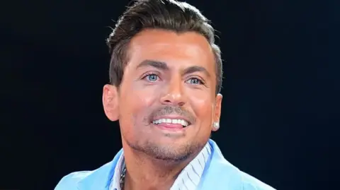 Paul Danan smiling in a light blue suit. He has dark brown hair, which is longer on top than it is on the side. He is wearing a large earring in one ear. 
